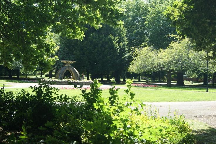 A stone monument in a park, with a lamppost and bin on the right and some people walking through the park.